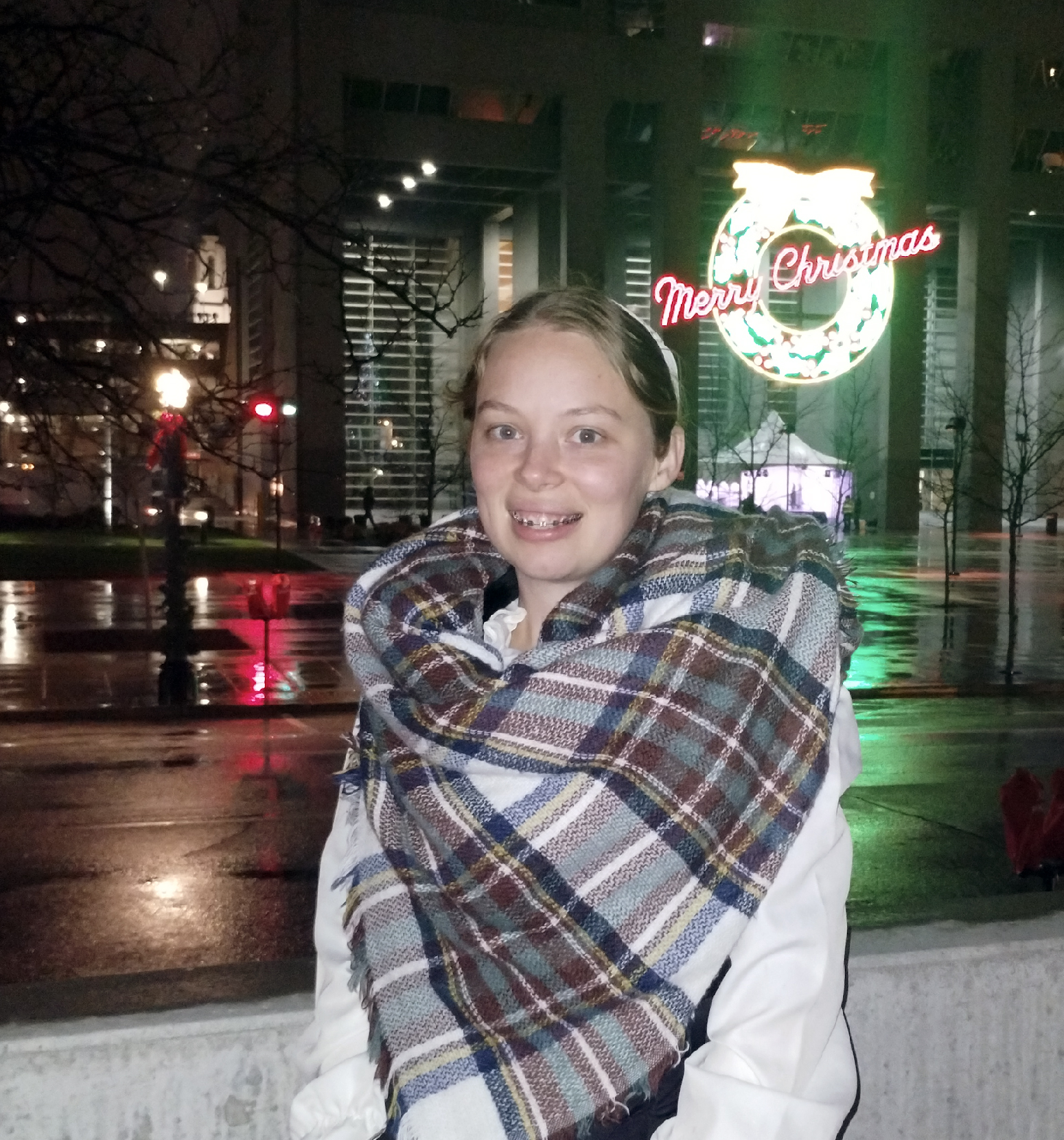 Image: TLA Graduate Rachel wrapped in a scarf in Downtown Fort Wayne, in front of a large wreath made of lights.